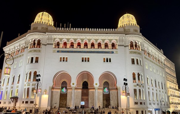 A beautifully illuminated historical building (Grand Post Office, Algier, Algeria) at night, featuring ornate architectural details, arched entranceways, and decorative domes. People gather at the base, enjoying the ambiance.