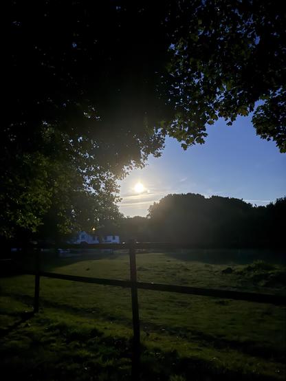 A serene evening scene featuring the sun setting behind trees, casting soft light in a lush green field. A wooden fence is visible in the foreground, with a house slightly illuminated in the background.